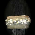 Un pain de mousse oasis de grande dimension décoré de fleurons d'orchidées et de galets oasis est posé sur  un vase noir.       