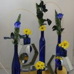 Jeu de bouteilles bleues, garnies de roses bleues et de chrysanthèmes jaunes, reliées par du rotin.