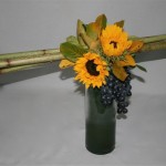 Un fagot de tiges, installé sur un vase haut et étroit, est décoré de fleurs de saison et de raisins.