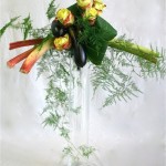 Sur un tube en verre, roses, aubergines et rhubarbe, sont accompagnées de feuilles de cordyline.