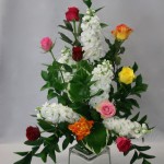 Bouquet triangulaire structuré de branches de ruscus et de giroflées, et animé de quelques roses multicolores.